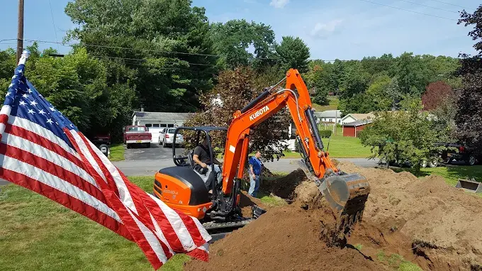 Justin's orange Kubota mini-excavator digging in a yard beside a large American flag